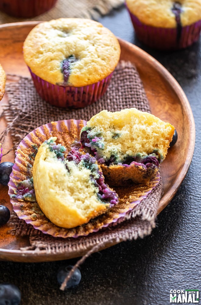 two blueberry muffins placed on a wooden plate with one of them cut in half to show the texture of the muffins