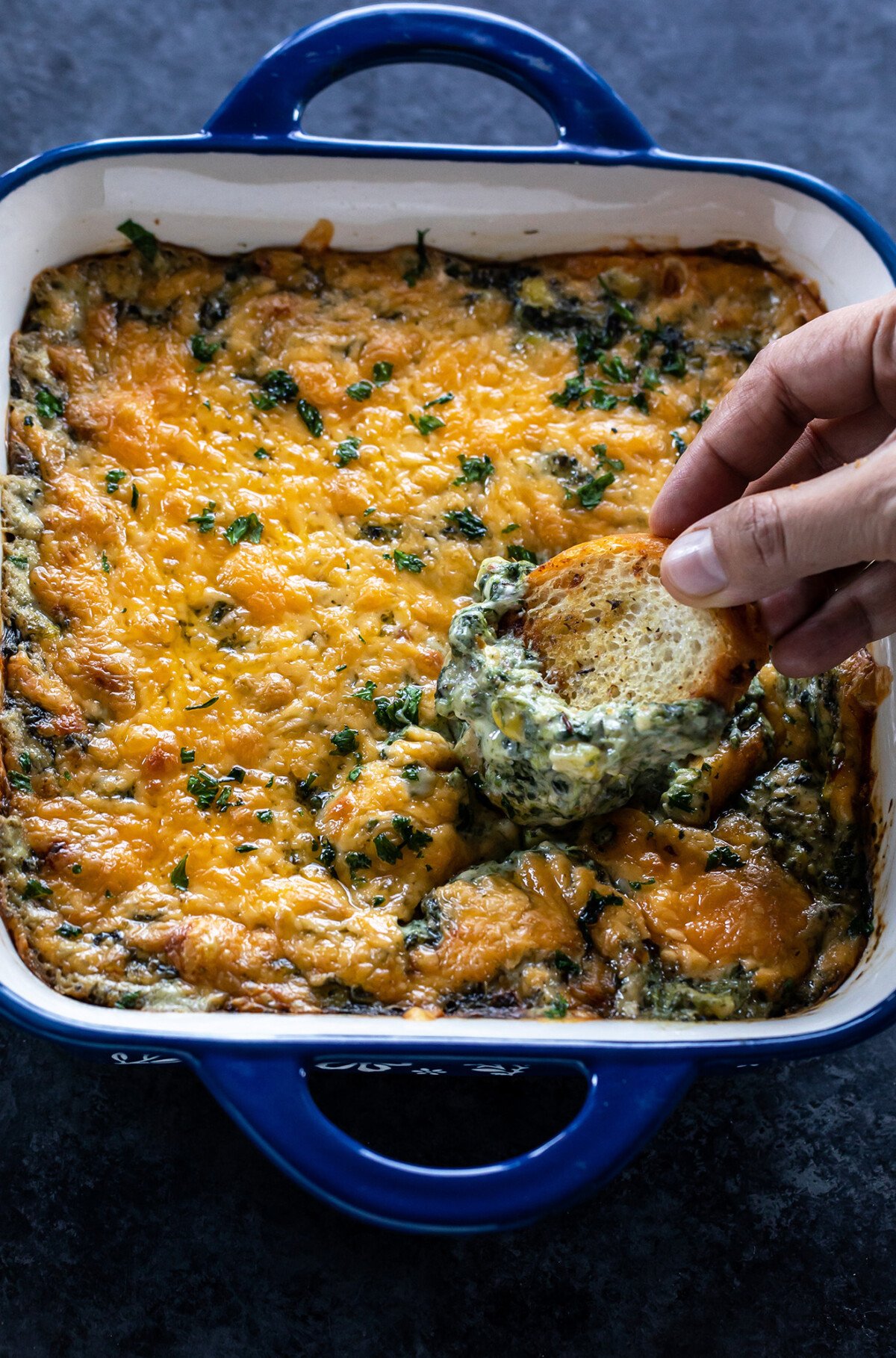 a hand holding a small pieces of bread digging into a cheesy dip
