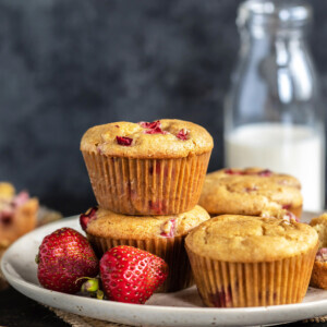 2 strawberry muffins stacked together with 2 more placed on the side and fresh strawberries placed in the front of the plate