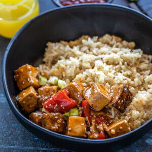 bowl of tofu topped with sesame seeds and brown rice with a glass of pineapple juice in the background with a bowl of hot sauce placed on the side