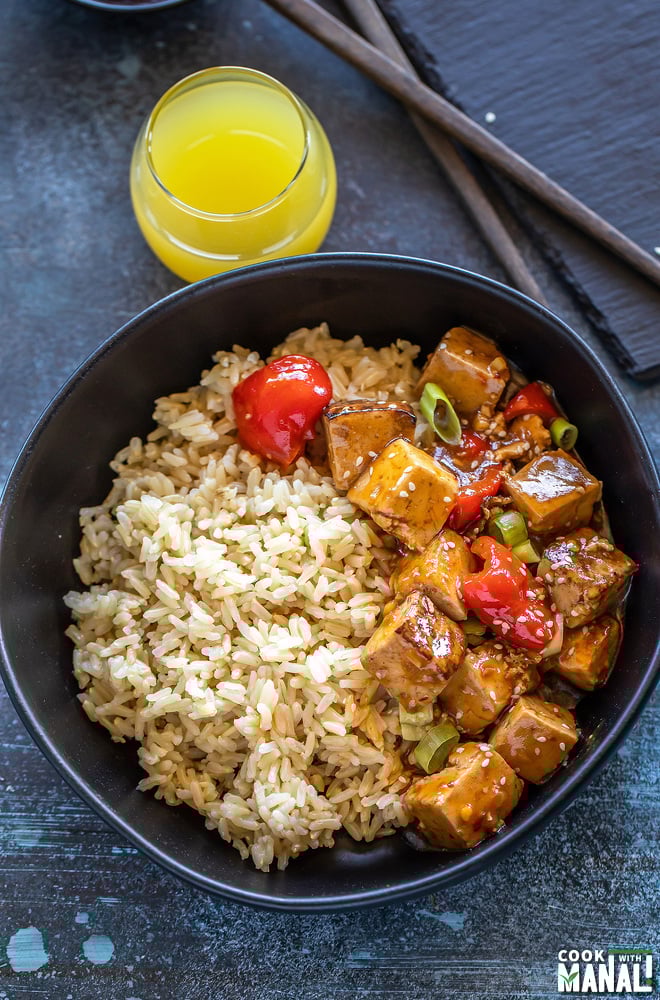 bowl of tofu topped with sesame seeds and brown rice with a glass of pineapple juice in the background with a bowl of hot sauce placed on the side