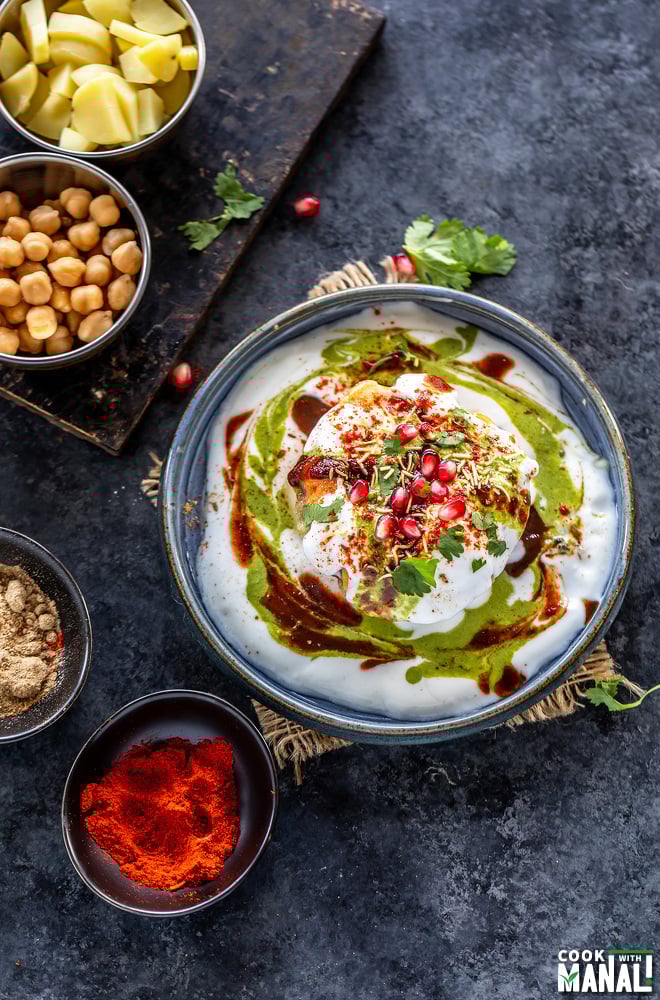 raj kachori placed in a plate garnished with pomegranate, cilantro and bowls of potatoes, chickpeas and spices placed on the side and background