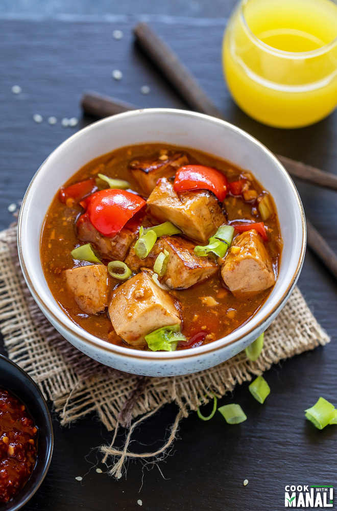 tofu and bell peppers in a white bowl garnished with green onion and a pair chopsticks on the side and a glass of pineapple juice on the side
