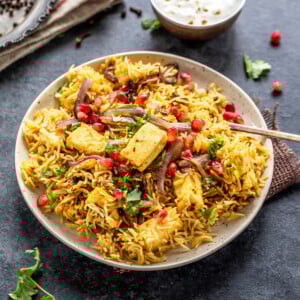 a plate full of rice and paneer, garnished with cilantro and pomegranate arils with a bowl of yogurt place on one side and a plate of spices placed on the other side