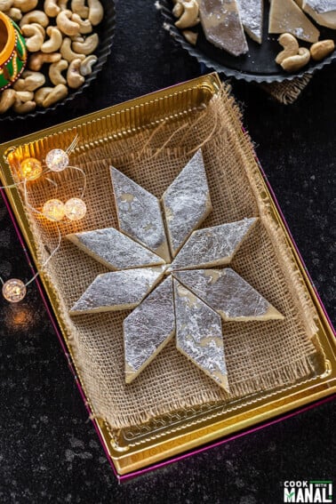 kaju katli arranged in a sweet box with small lights on the side and plate of cashews in the background