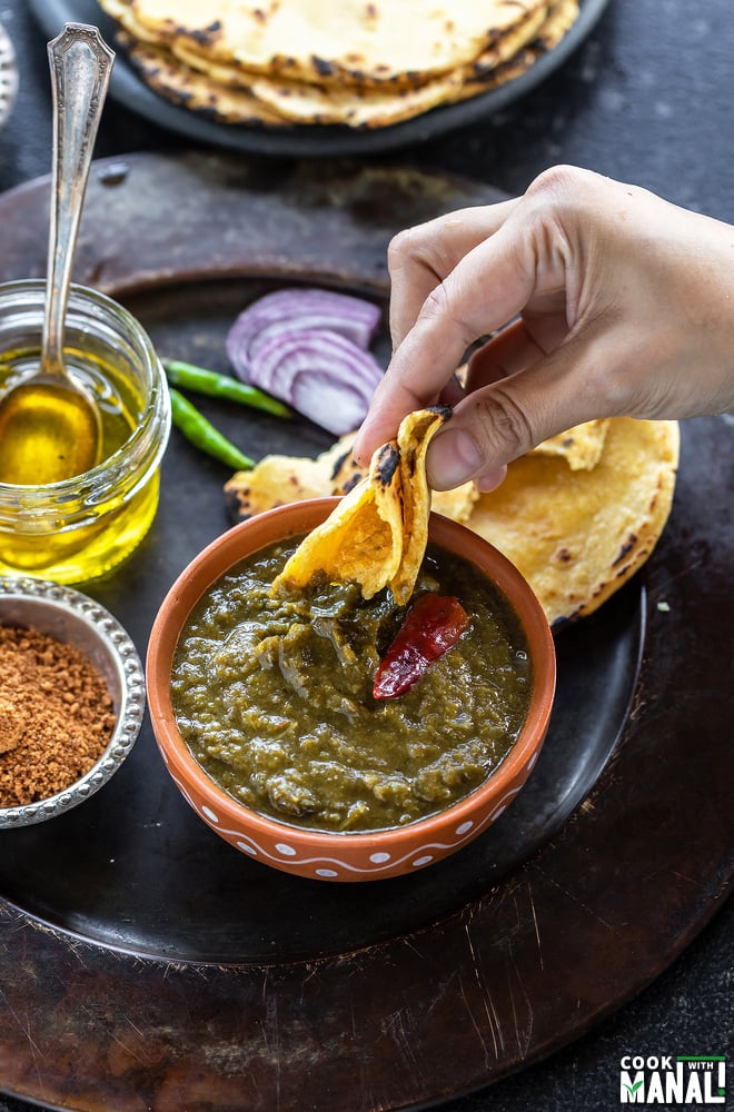 a hand dipping a bite from the makki ki roti into a bowl of saag