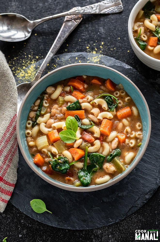 bowl of minestrone soup with two spoons in the background and a kitchen towel on the side