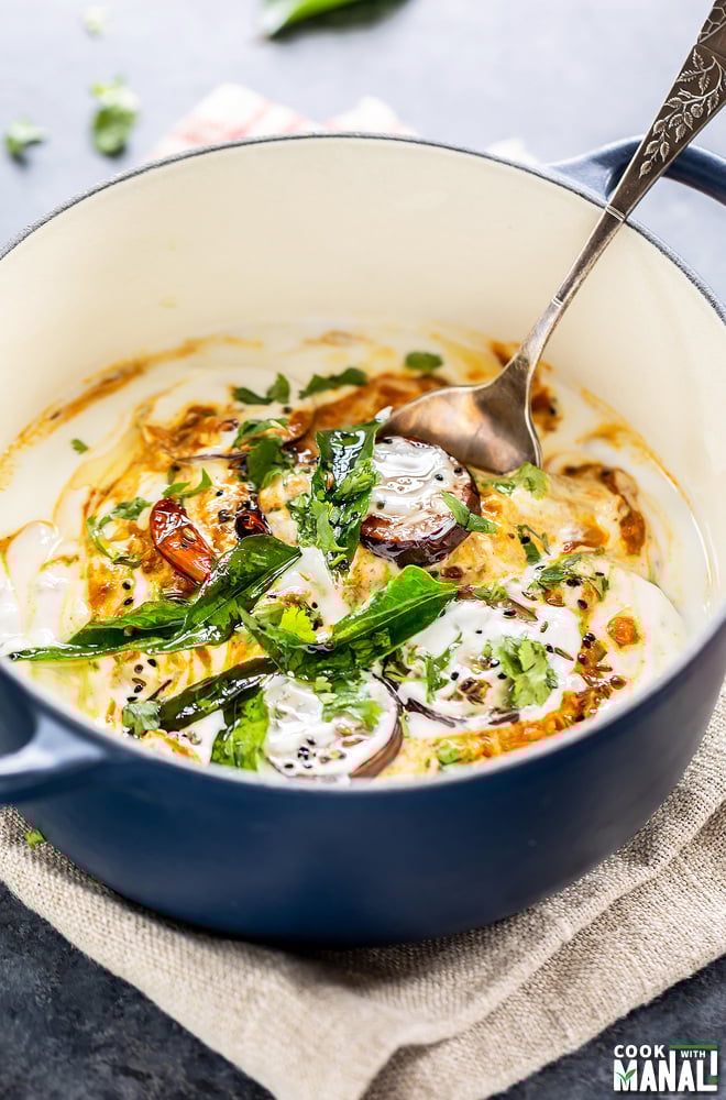 eggplant and yogurt dish served in a dutch oven with a spoon and garnished with cilantro, curry leaves