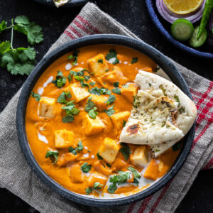 bowl of paneer butter masala served with a piece of naan and garnished with cilantro and cream