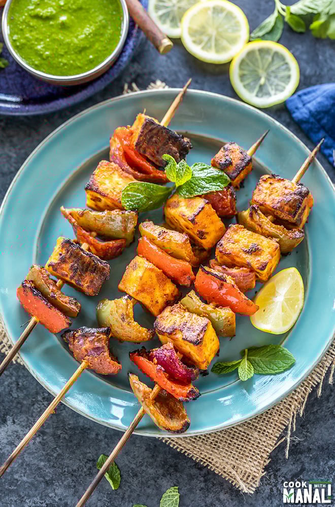 paneer tikka skewers arranged on a blue plate with lemon wedge on the side and bowl of cilantro chutney in the background