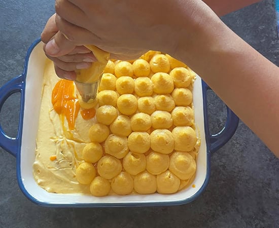 a hand piping the top of a mango tiramisu using a round piping tip