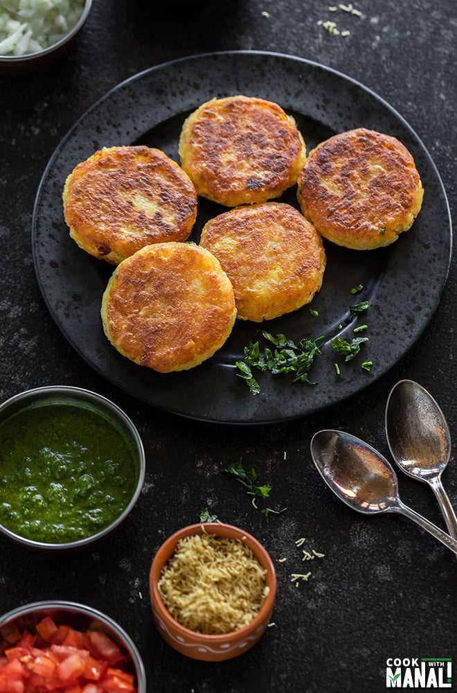 plate of 5 potato patties in a black plate surrounded by several bowls of chutney and sev