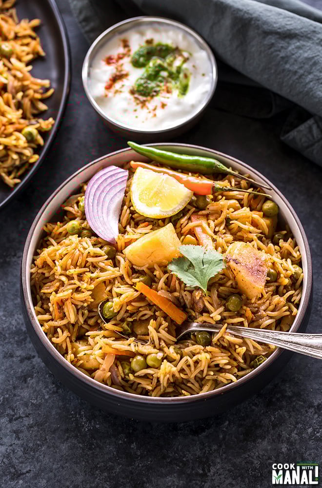 overhead shot of vegetable biryani in a black bowl garnished with lemon wedge, sliced onion and green chili