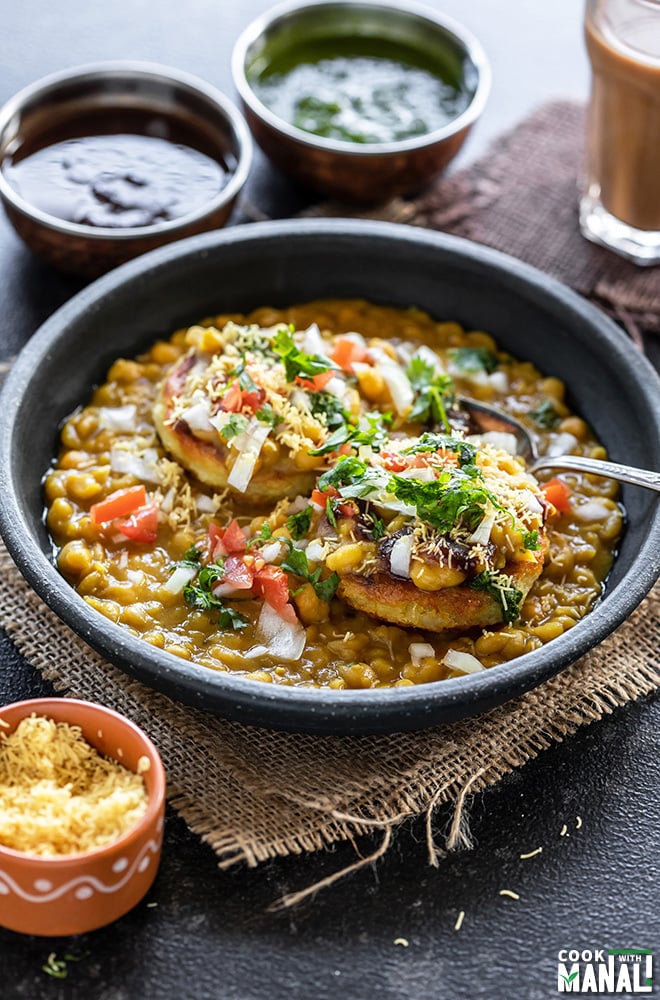 ragda patties served in a black bowl with a spoon and glass of chai and several bowls of chutney in the background and on the sides