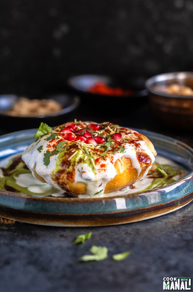 raj kachori placed on a plate topped with yogurt, chutney, pomegranate, cilantro and bowl of spices placed in the background