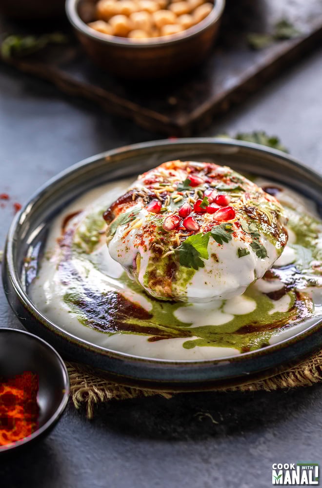 raj kachori placed on a plate topped with yogurt, chutney, pomegranate, cilantro and bowl of spices placed in the background