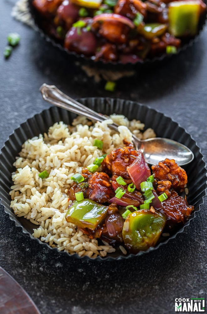 chilli paneer served with brown rice and a spoon in a black rimmed plate