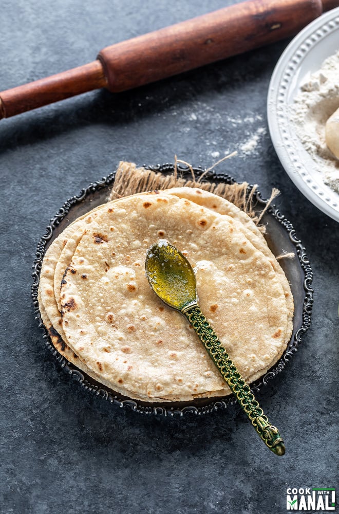 a golden color spoon placed on a stack of roti served in a silver plate with a rolling pin in the background