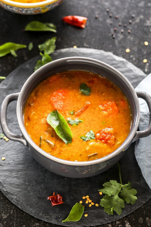 sambar served in a grey bowl garnished with curry leaves with twigs on cilantro and more curry leaves scattered all around