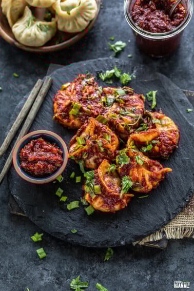 momos topped with cilantro, green onions arranged on a black board with a bowl of hot sauce and pair of chopsticks placed on the side