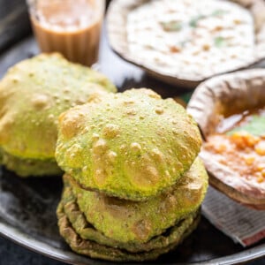 stack of puris placed in a plate along with glass of chai and bowls of curry and raita