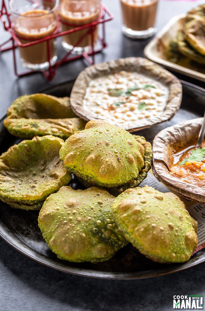 5 spinach puri in a round plate with bowls of potato curry and boondi raita on the side and glasses of chai in the background