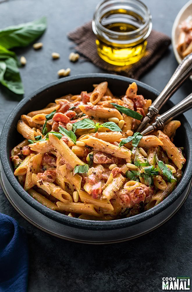 tomato basil pasta in a black bowl with 2 forks. There are also some basil leaves and a small jar of olive oil in the background