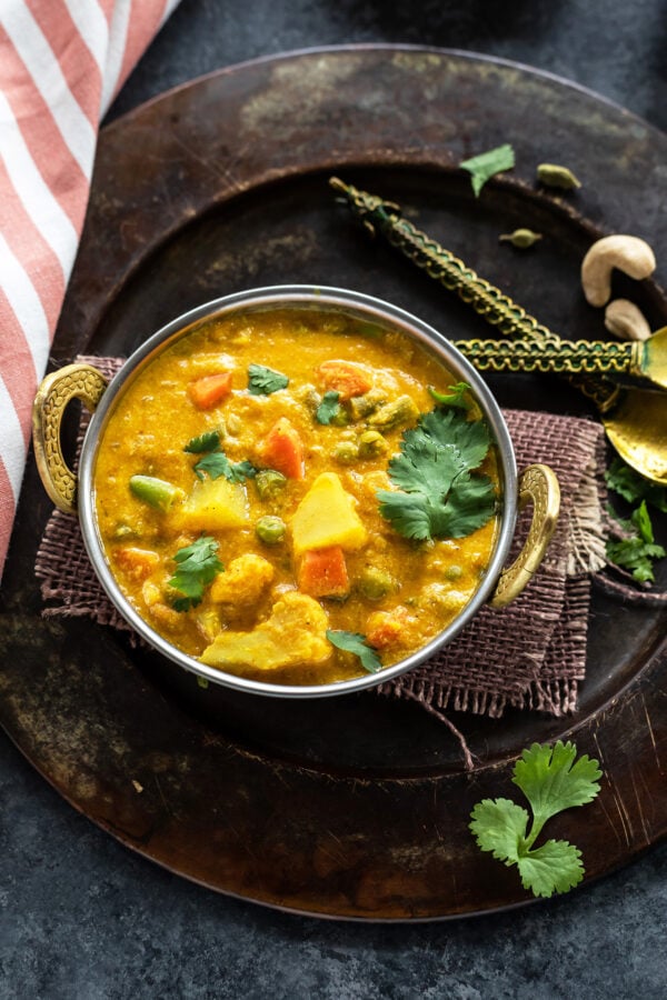 overhead shot of veg kurma served in a copper kadai with a kitchen towel placed on one side and two golden color antique spoon on the other side