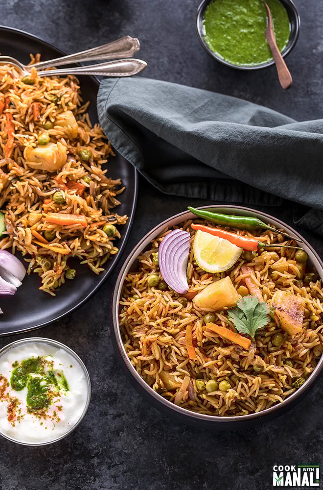 vegetable biryani in a black bowl with a bowl of yogurt on the side