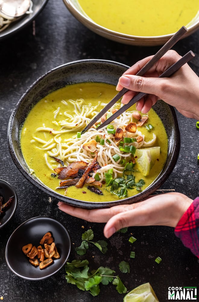 pair of hand holding a bowl of noodle soup from one side and holding chopsticks with the other hand